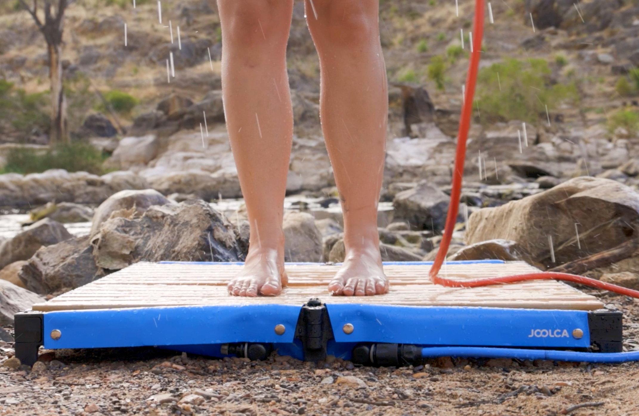 Bare feet standing on a blue portable outdoor shower base on rocky ground with water droplets falling, surrounded by natural outdoor scenery.