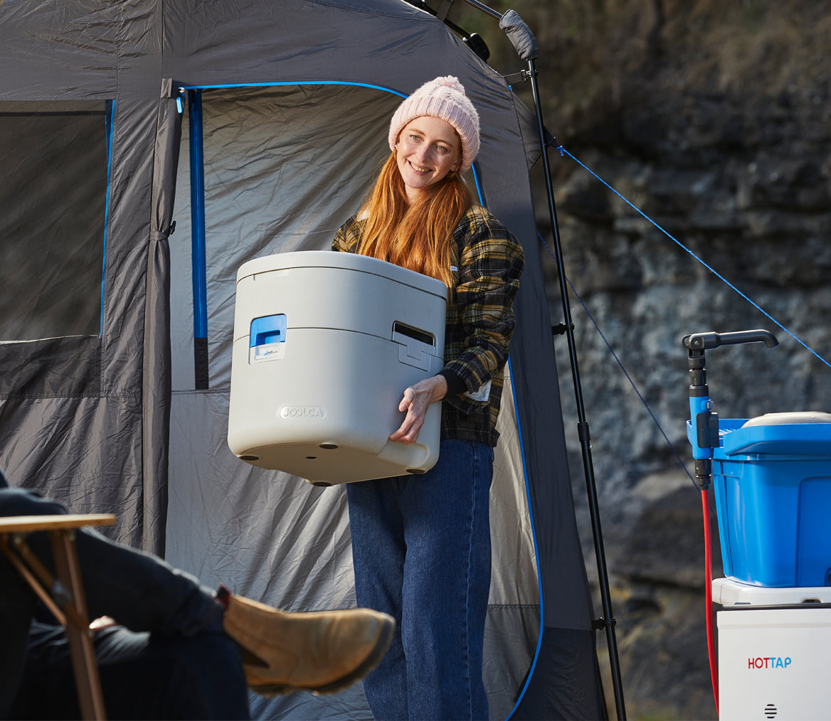 Woman wearing a beanie and plaid shirt holds a portable composting toilet outside a privacy tent with camping equipment and rocky terrain in the background.
