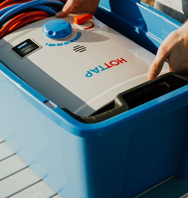 Hands placing a portable tankless water heater labeled "HOTTAP" inside a blue carrying case with coiled red and blue hoses visible in the background.
