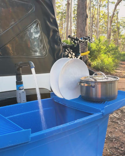 Water running from the HOTTAP V2 tap into the blue Flexi Sink beside a camper trailer, with plates and a pot draining alongside.