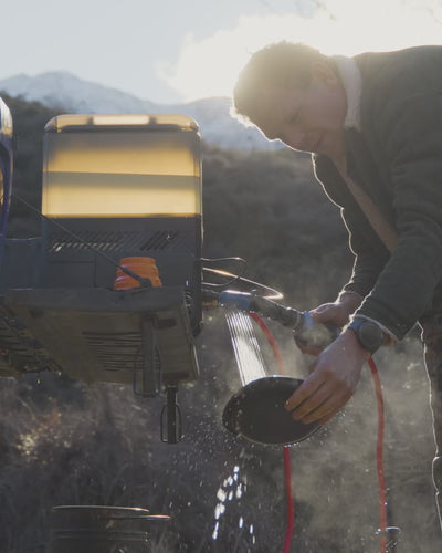 Camper washing a pan with a brush over the blue Joolca Flexi Sink outdoors, rinsing under a HOTTAP water heater spray.