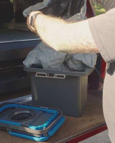 Man at a vehicle tailgate lifting the lined bag out of the GottaGo solids cassette, the blue lid frame set down alongside.