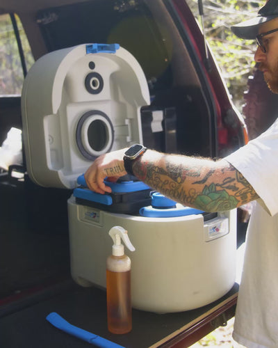 Man lifting the upper bowl away at a vehicle tailgate to reach the two blue-capped GottaGo cassettes underneath.